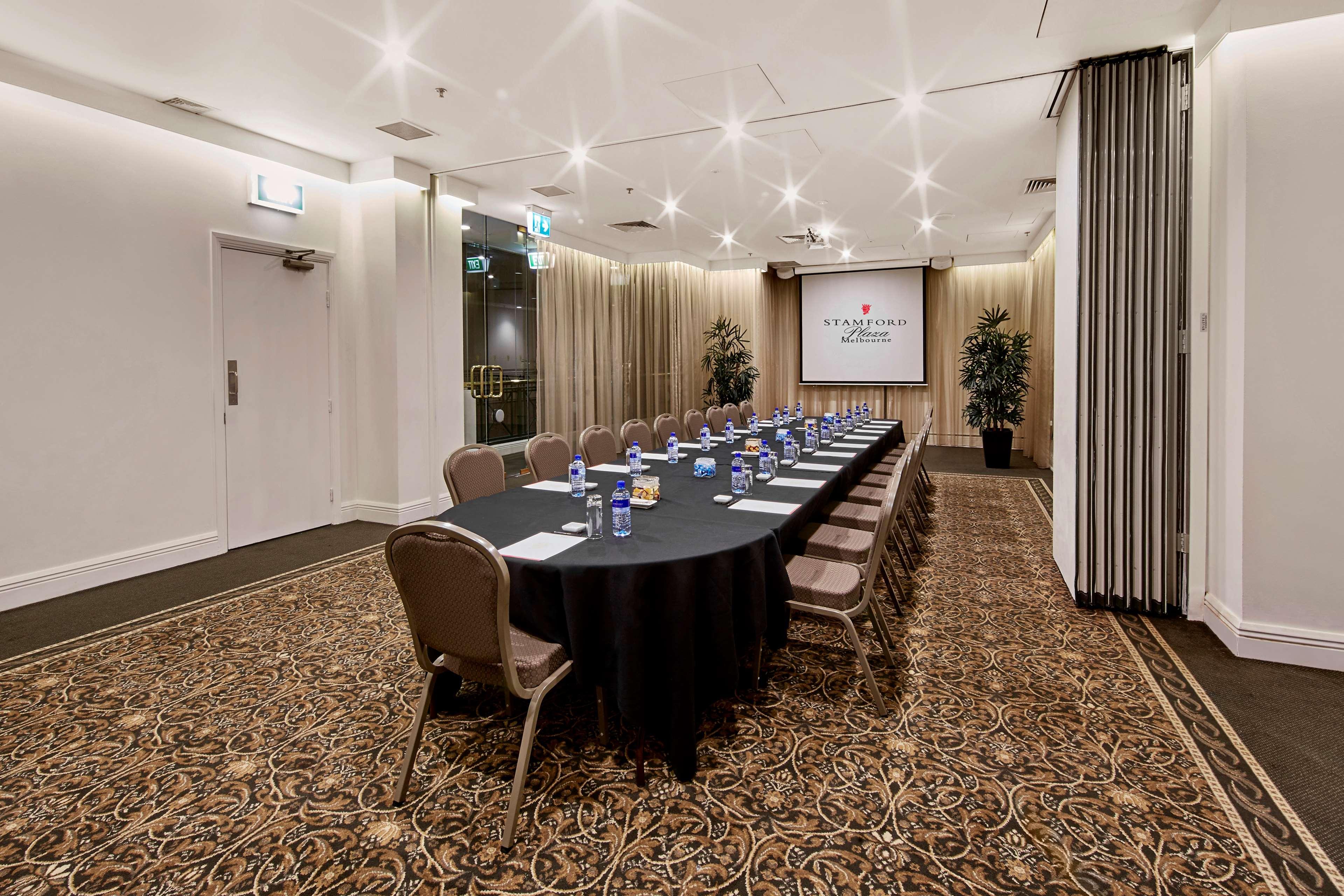 Hotel Stamford Plaza Melbourne Zewnętrze zdjęcie The photo shows a meeting room set up for a conference or presentation. There is a long, rectangular table covered with a black tablecloth, surrounded by chairs. Each seat has a water bottle placed in front of it. At the front of the room, there is a