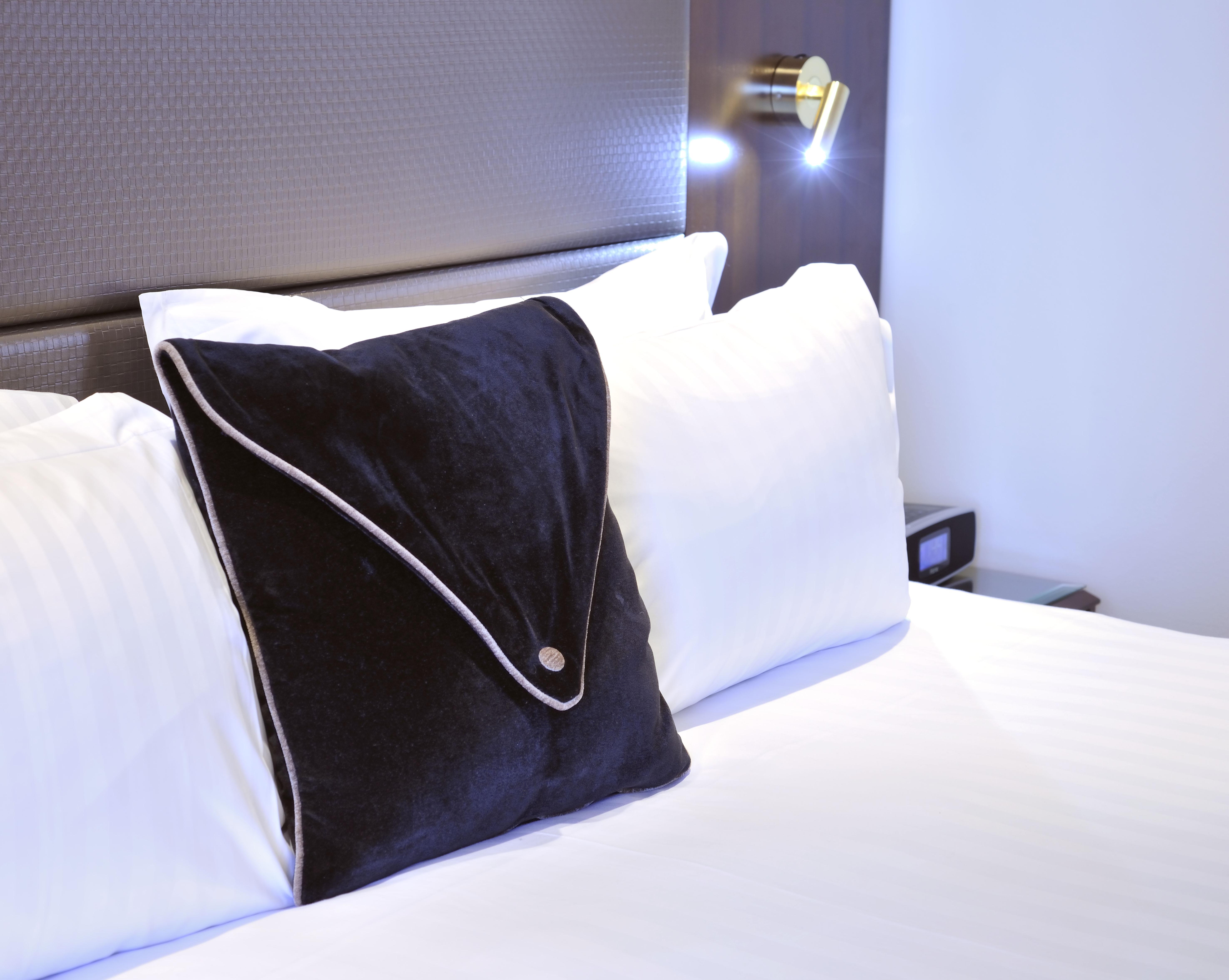 Hotel Stamford Plaza Melbourne Zewnętrze zdjęcie The photo shows a neatly arranged bed with white linens. There are several pillows, with one black pillow featuring a distinctive design on top of the white pillows. The headboard of the bed is visible, along with a stylish wall-mounted light fixture
