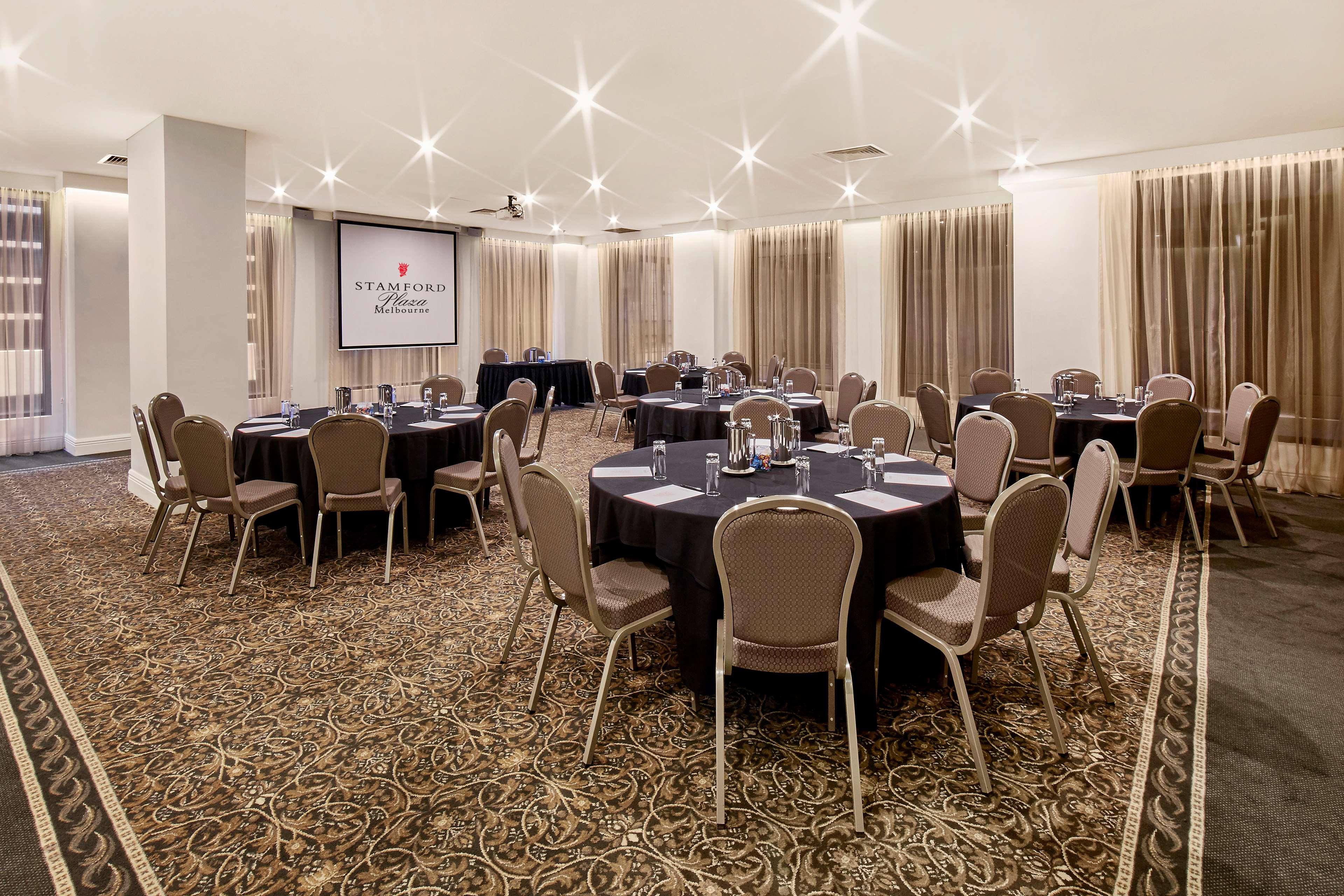 Hotel Stamford Plaza Melbourne Zewnętrze zdjęcie The photo shows a well-lit event space set up for a gathering or meeting. There are several round tables covered with black tablecloths, each surrounded by beige chairs. In the background, a large screen displays a logo or branding, likely related to