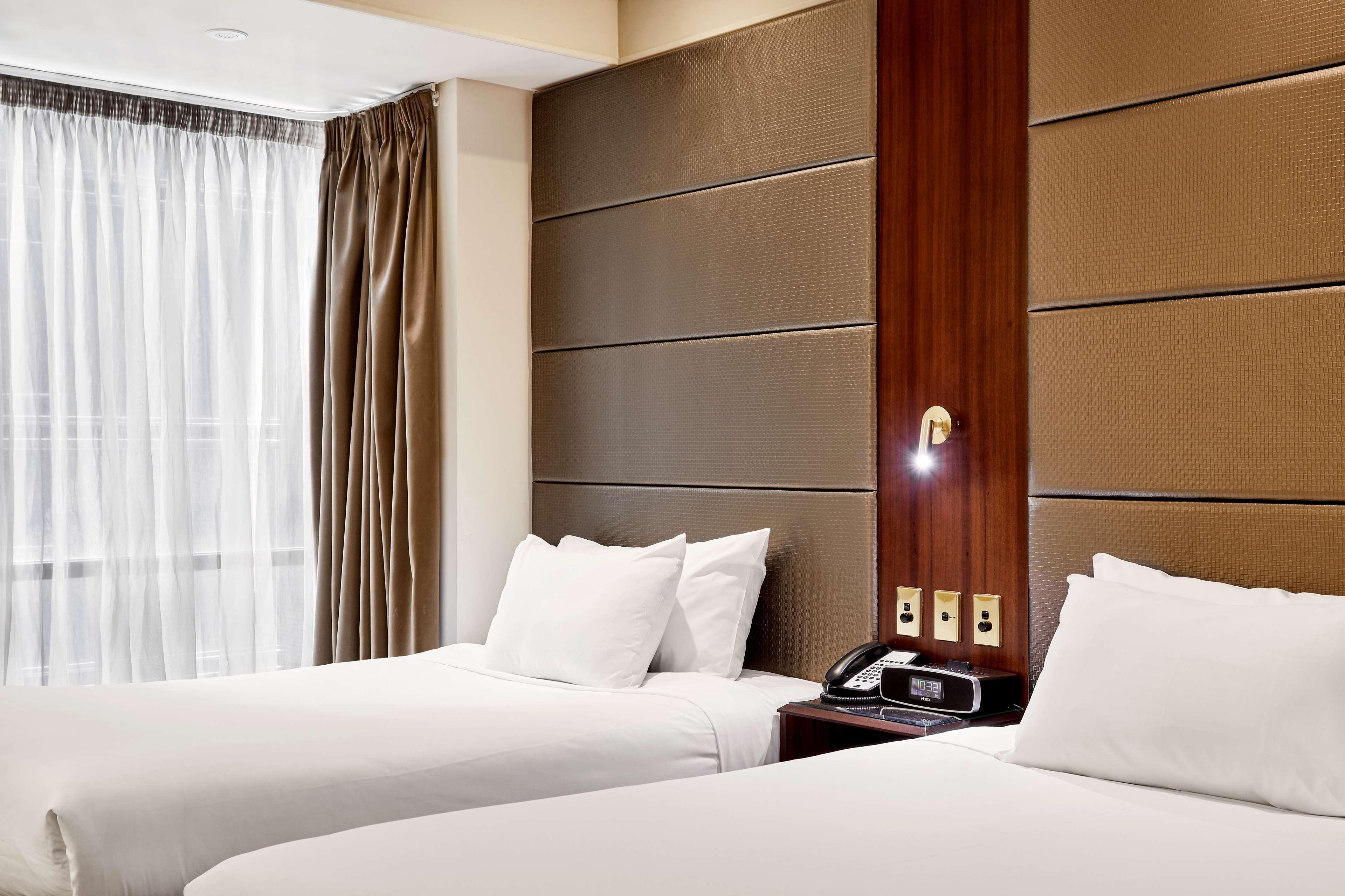 Hotel Stamford Plaza Melbourne Zewnętrze zdjęcie The photo shows a modern hotel room featuring two twin beds with white linens and fluffy pillows. The walls behind the beds have a combination of textured panels in neutral colors. There is a bedside table with a lamp and a phone. A large window with