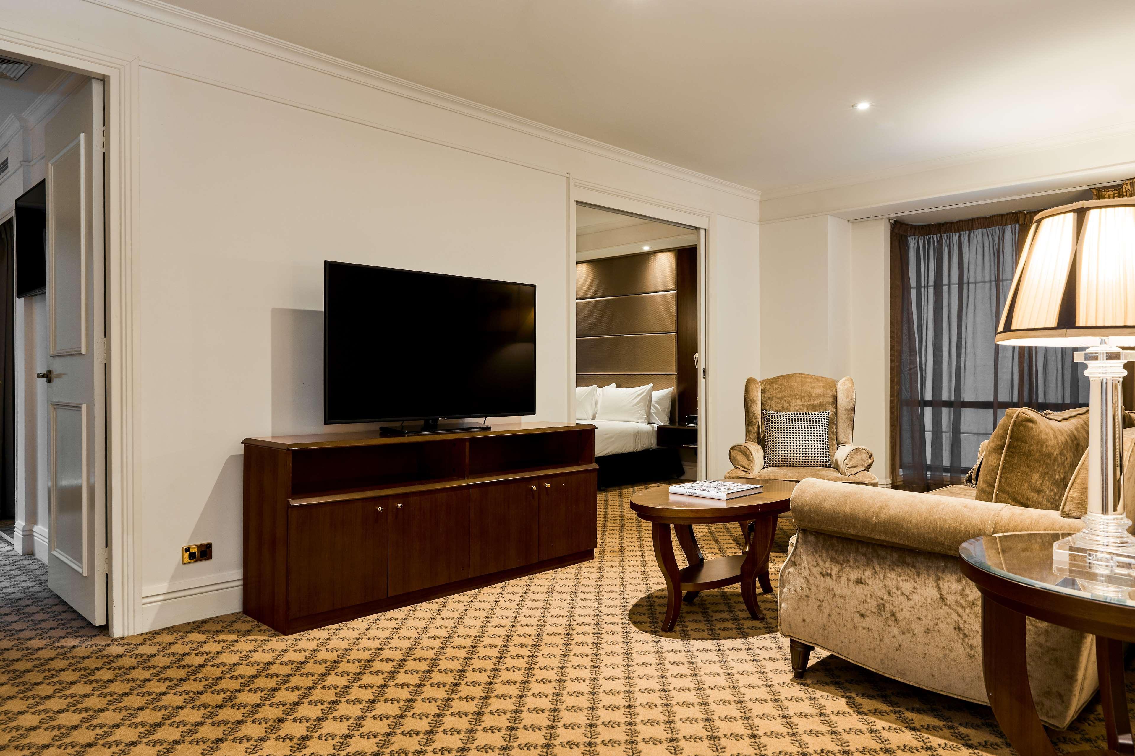 Hotel Stamford Plaza Melbourne Zewnętrze zdjęcie The photo shows a well-furnished room, likely a hotel suite or a living area. There is a large flat-screen television mounted on a wooden entertainment unit. In front of the TV, there is a round wooden coffee table. To the side, a comfortable armchai
