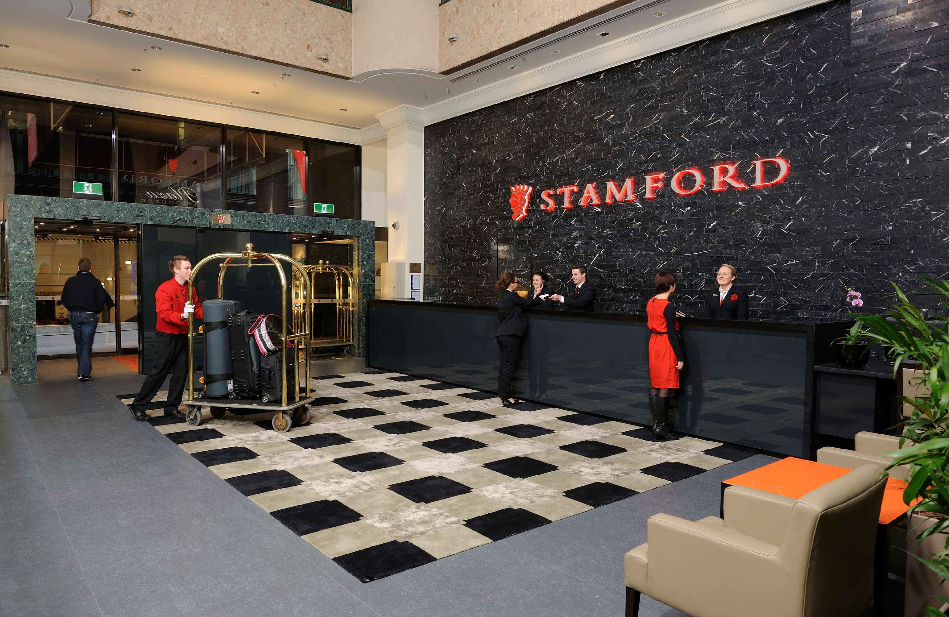 Hotel Stamford Plaza Melbourne Wnętrze zdjęcie The photo shows the interior of a hotel lobby, specifically the Stamford Hotel. There is a large reception desk where several staff members are attending to guests. In the foreground, a bell cart loaded with luggage is positioned beside the reception