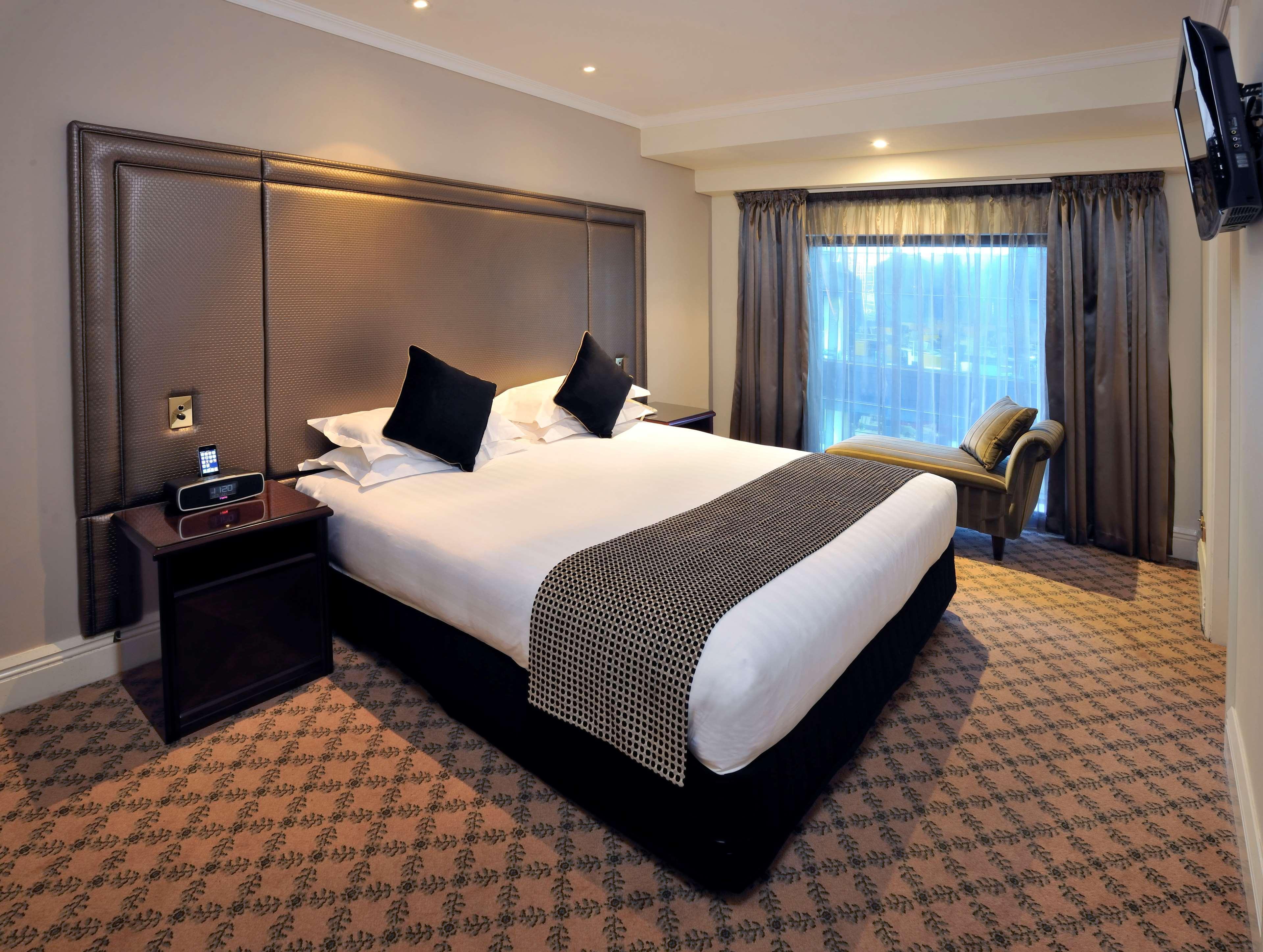 Hotel Stamford Plaza Melbourne Zewnętrze zdjęcie The photo shows a hotel room featuring a neatly made king-sized bed with dark pillows. The headboard is upholstered in a dark material, and there is a decorative blanket at the foot of the bed. Beside the bed are two nightstands, each equipped with l