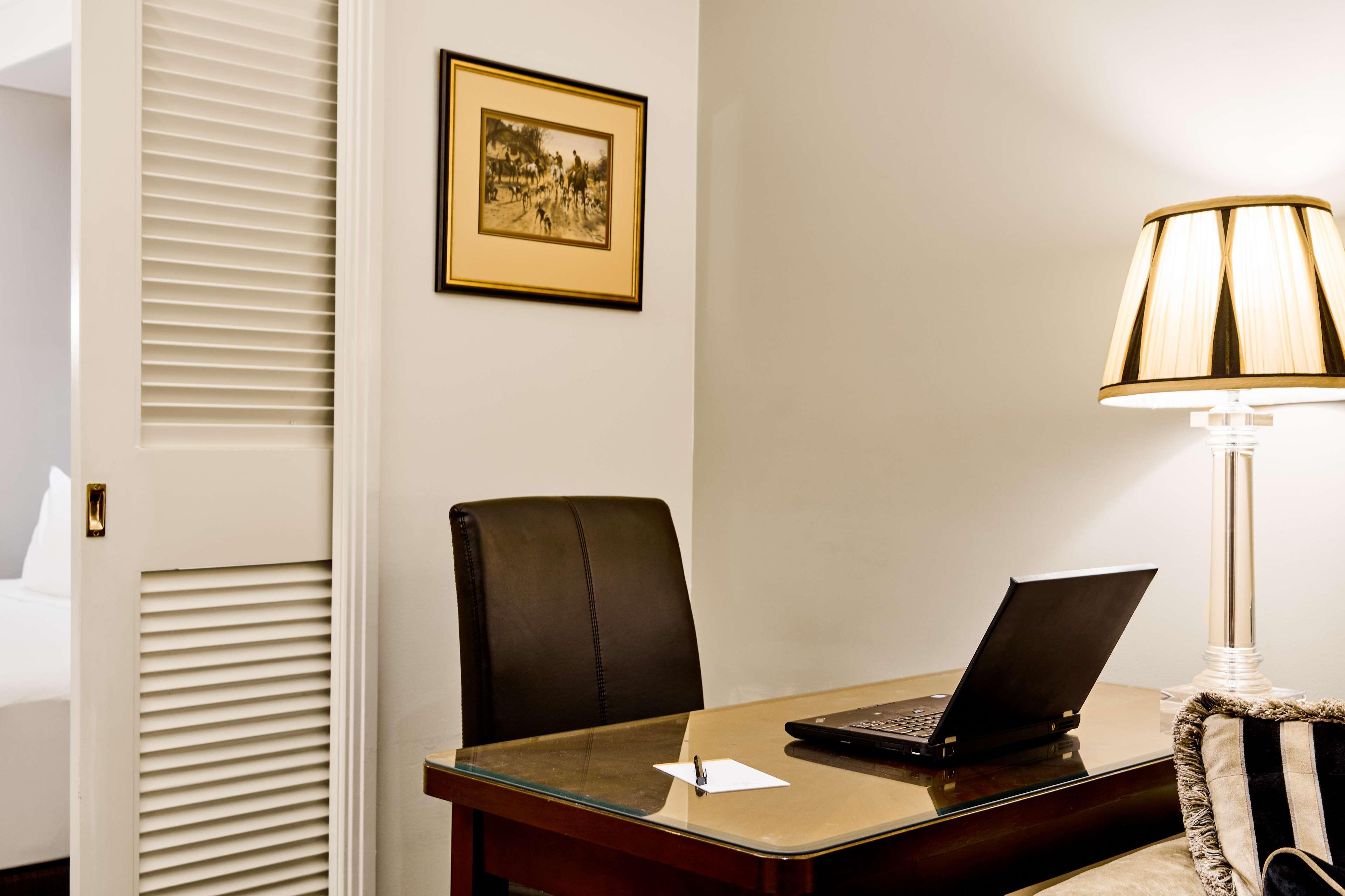 Hotel Stamford Plaza Melbourne Zewnętrze zdjęcie The photo shows a small office or workspace. There is a desk with a glass surface, and on it sits a laptop computer. A piece of paper or notecard is also placed on the desk. Behind the desk is a black office chair. A wall lamp with a striped shade is