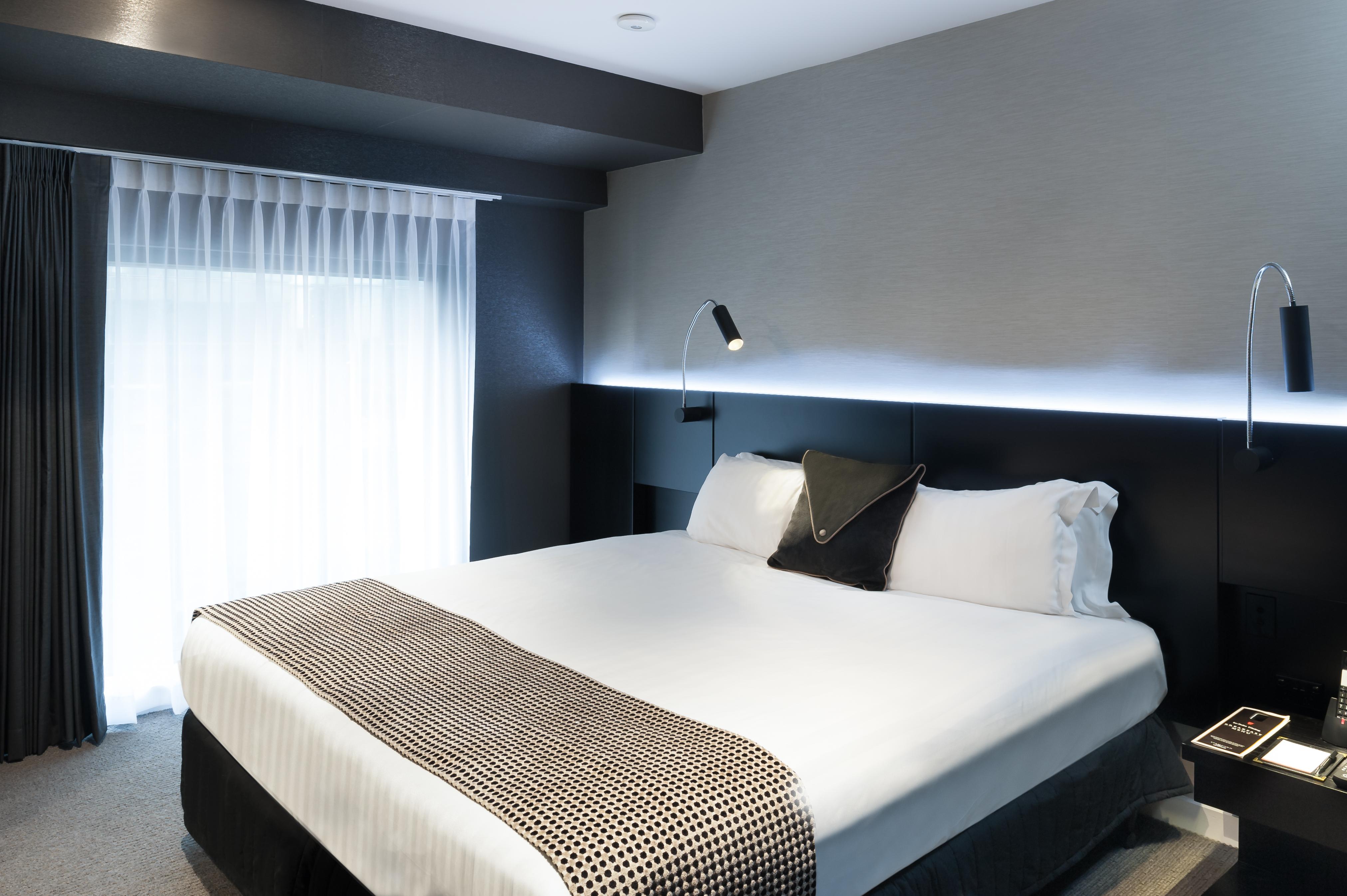 Hotel Stamford Plaza Melbourne Zewnętrze zdjęcie The photo shows a modern hotel room featuring a large bed with white linens and a decorative throw blanket. The room has a sleek and contemporary design, with gray walls and a soft lighting strip along the headboard. A window is adorned with sheer cu
