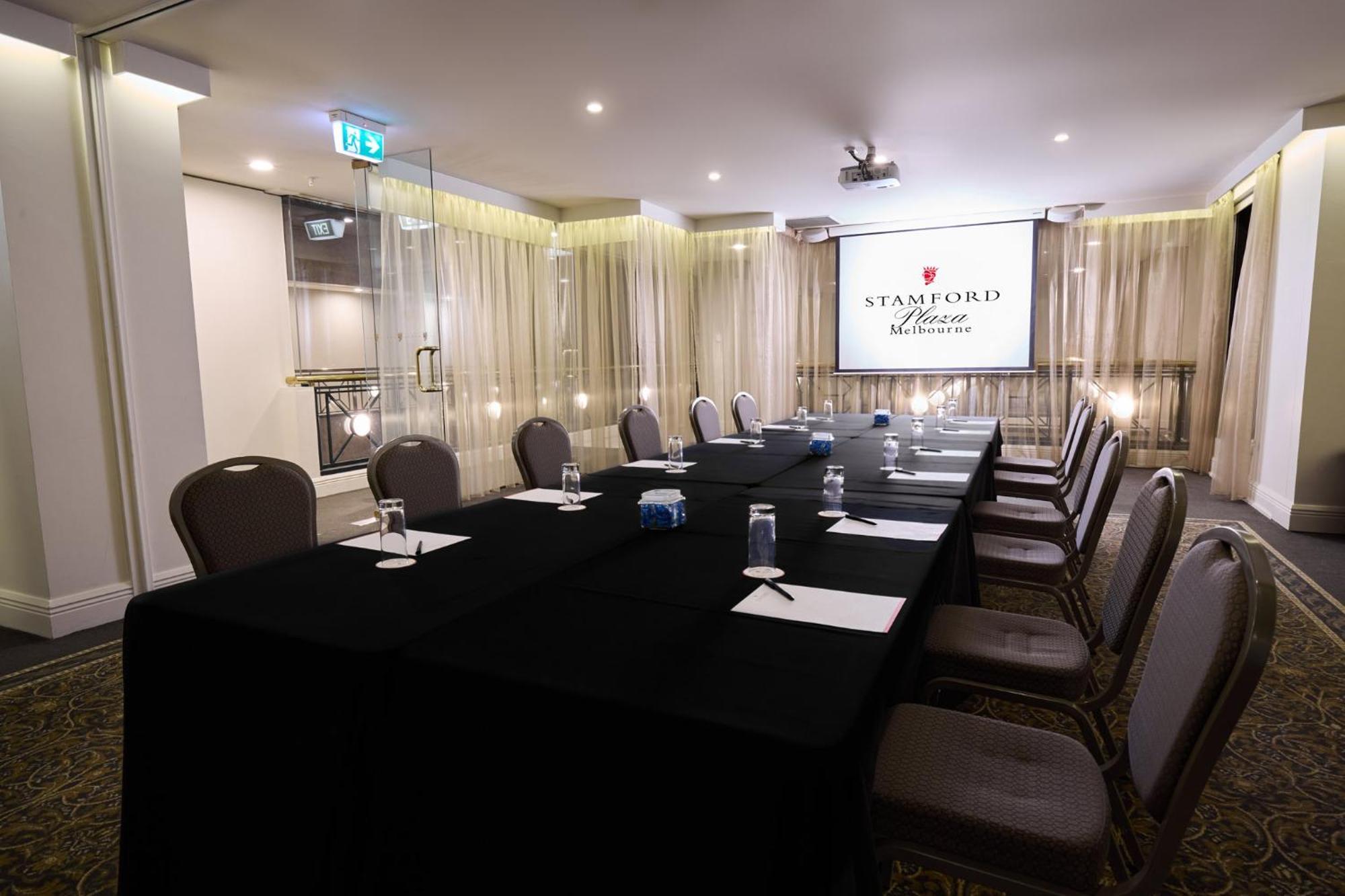 Hotel Stamford Plaza Melbourne Zewnętrze zdjęcie The photo shows a well-appointed conference room designed for meetings or presentations. There is a long rectangular table covered with a black tablecloth, surrounded by upholstered chairs. On the table, there are several folded pieces of paper and g