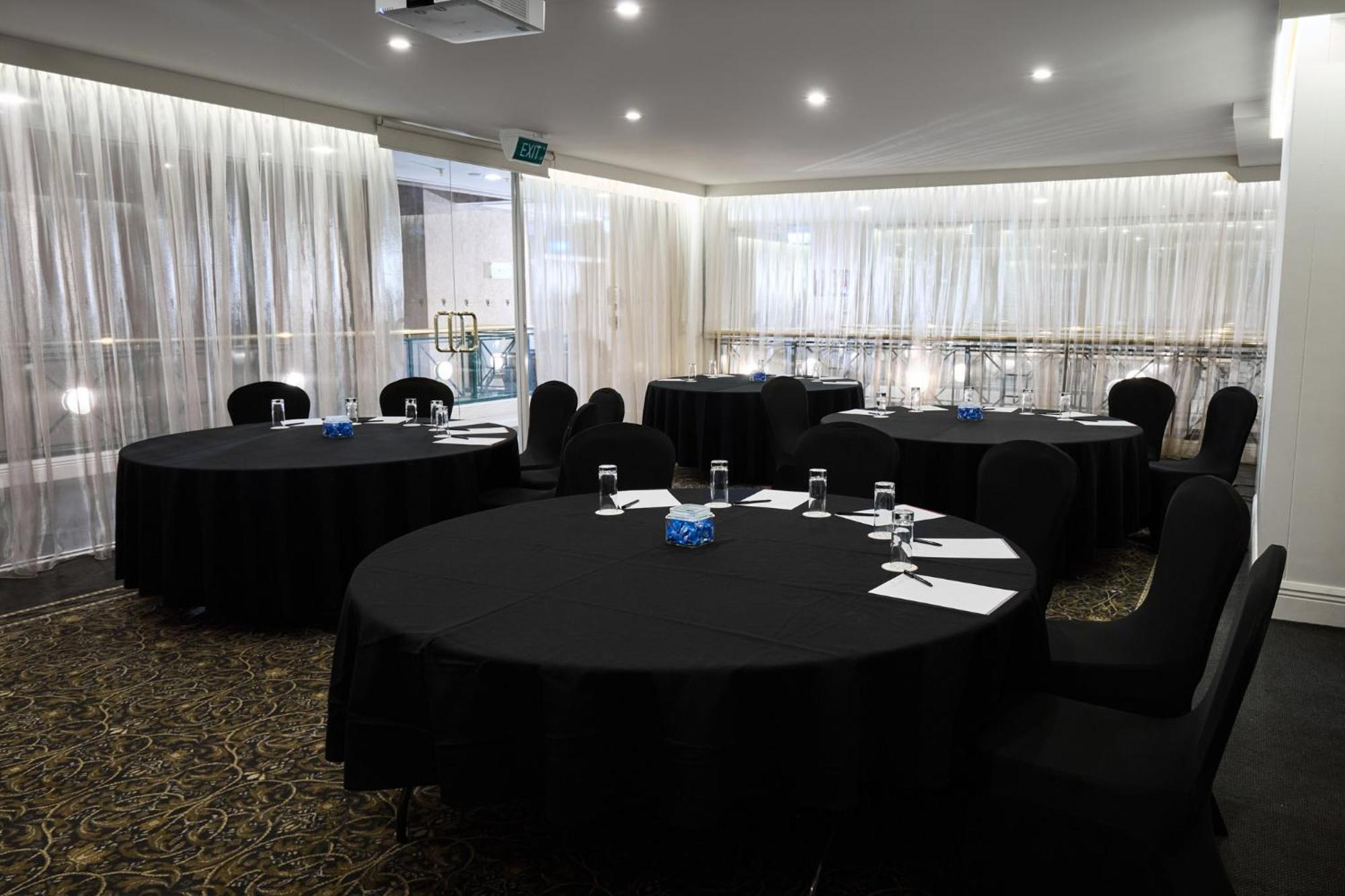 Hotel Stamford Plaza Melbourne Zewnętrze zdjęcie The photo shows a meeting or conference room set up with several round tables covered in black tablecloths. Each table is accompanied by black chairs. There are some white notepads and pens placed on the tables, and a small decorative item, possibly 