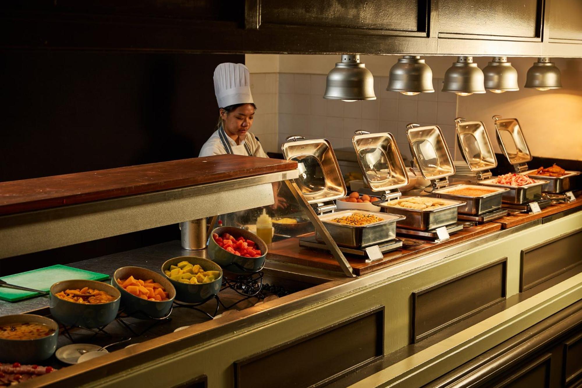 Hotel Stamford Plaza Melbourne Zewnętrze zdjęcie The photo shows a buffet setup in a dining area. There is a chef in a white uniform and hat, standing behind a counter filled with various food options. The buffet features several metal serving trays containing different dishes, with warming lights 