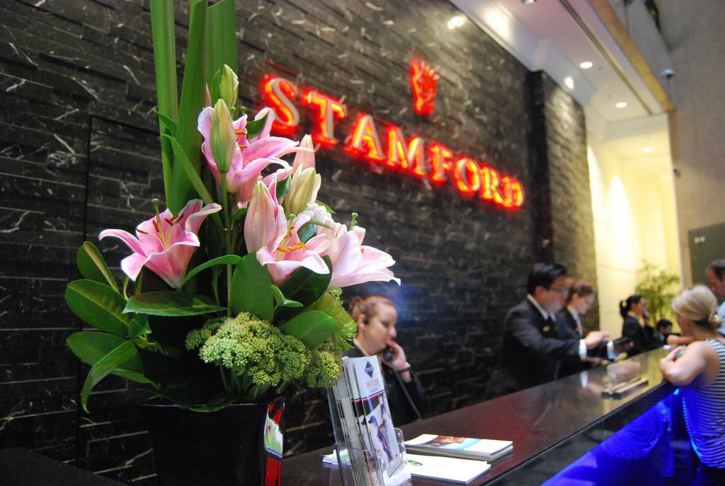 Hotel Stamford Plaza Melbourne Zewnętrze zdjęcie The photo shows a hotel reception area with a prominent sign that reads "STAMFORD" in bright red letters. In the foreground, there is a decorative arrangement of flowers, primarily featuring pink lilies, along with greenery. The reception counter is 